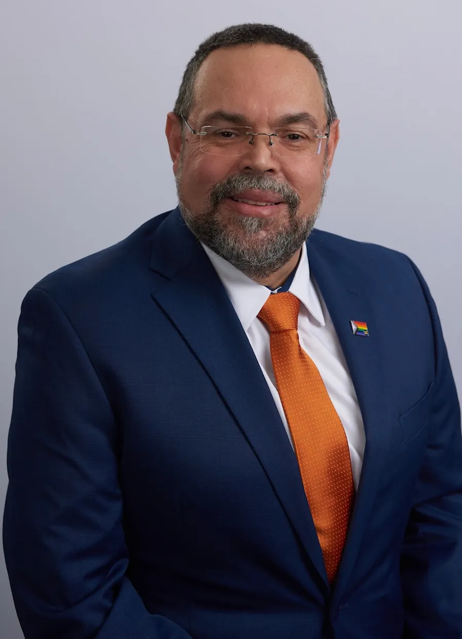 Studio headshot of Wilfredo in a navy suit with an orange tie and a Progress Pride pin